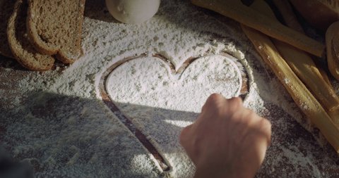 Cinematic close up shot of professional artisan baker chef is drawing heart shape with flour on wooden board with bread, loaf and breadsticks on background in rustic bakery kitchen.