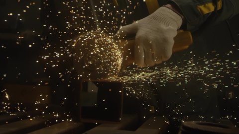Industrial worker using an angle grinder and cutting a metal square pipe. Locksmith in special clothes in production. Industry machinery plant interior. Manufacturing metal structures. Close up, 4K
