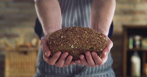 Cinematic close up shot of professional artisan baker is showing in camera just prepared fresh rye whole grains brown bread taken out of oven in rustic bakery kitchen.