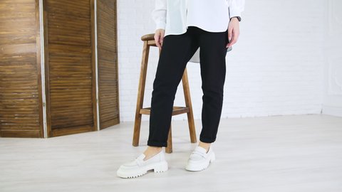 Lady in white shirt and black jeans demonstrates white fashionable shoes. Woman walks in plain loafers with massive soles back and forth near wooden tabouret.