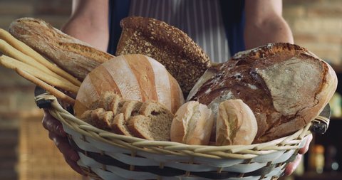 Cinematic close up shot of professional artisan baker presenting in camera basket with assortment of variety crunchy just prepared fresh bread and baked buns in rustic bakery kitchen.