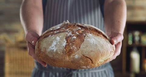 Cinematic macro shot of professional artisan baker is showing in camera just prepared fresh whole grains white bread taken out of oven in rustic bakery kitchen.