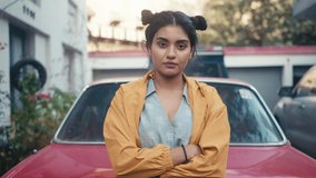 A young modern stylish retro smiling Indian Asian woman or girl is standing outdoors leaning on red vintage car and looking at camera in a city setting. Concept of youth, ambition, rebel, revolution