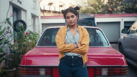 A young modern stylish retro smiling Indian Asian woman or girl is standing outdoors leaning on red vintage car and looking at camera in a city setting. Concept of youth, ambition, rebel, revolution
