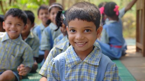 Close up shot of a cute smiling Indian Rural schoolgirl in uniform looking at the camera with a group of kids or fellow students sitting in the background, Kudal, Maharashtra, India (March 2020)