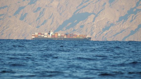 Cargo ship transporting goods off coast of Sinai. Freighter carries shipping containers near shore of Saudi Arabia. Merchant vessel, watercraft moving past rocky place. Shipment, reaching load by sea
