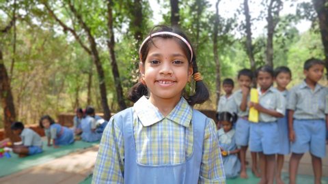 A cute smiling Indian Rural girl in uniform standing outdoors school premises and looking at the camera with a group of students in the background in a village, Kudal, Maharashtra, India (March 2022)