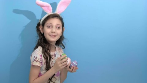 4k. Happy easter. Beautiful cute little girl in Easter bunny ears holding painted easter eggs on blue background going to celebrate Easter, stands over yellow studio background. Spring holiday concept