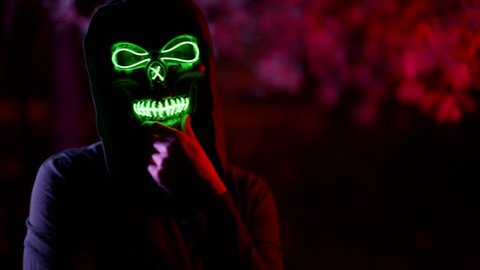 portrait of a person in a terrible mask of death and in a dark hood against a background of branches illuminated by red light. the character rubs chin and the bridge of nose, then beckons to him