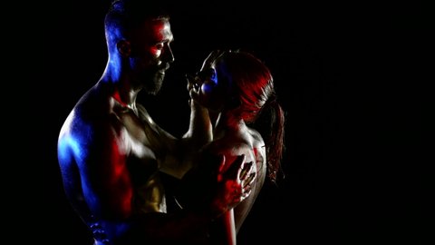 a couple with golden metallic skin hugs and touches each other on a black background. profile view. red, blue and white light. the dark key