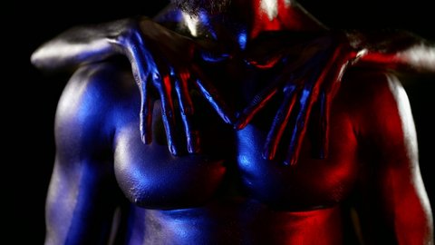 close-up of a muscular male torso with golden metallic skin. the woman stands behind the man and symmetrically slides hands over his skin. red, blue and white light. without faces dark key