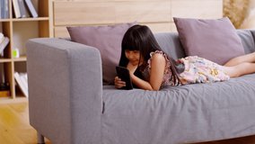 Cute little girl uses smartphone while prone at the sofa in the living room. Child surfing the internet on computer, browses through internet and watches cartoons online.