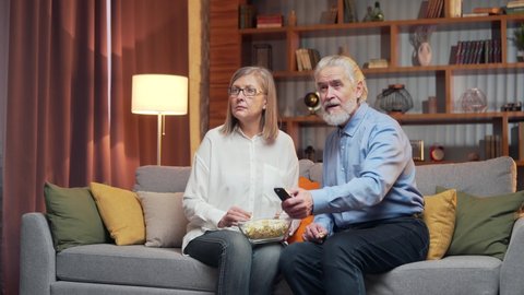 Bored Carefree grey-haired older couple at home choose a TV show to watch and switch channels with a TV remote control. family cannot choose a show. difficult choice, there's nothing to watch