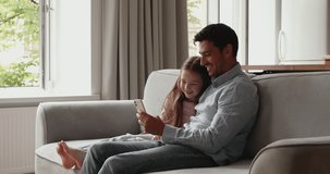 In cozy living room young father and little daughter look at smartphone screen watch vlog, use mobile app spend weekend together at home. Young generation and modern wireless tech overuse, fun concept