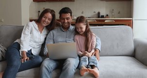 Couple and little 6s daughter gather in domestic room sit on cozy sofa enjoy weekend, use laptop watch family movie laughing feel happy. Young generation use modern tech for leisure at home concept