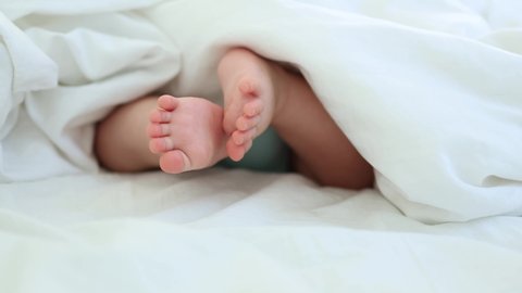close-up bare feet and toes of infant baby legs in white sheet. newborn tiny fingers feet moving. caucasian little baby foot macro view. barefoot child legs 