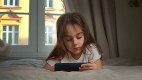 An excited little girl is lying on the bed and playing video games on her smartphone in a cozy room at home. A happy preschool girl who wins players and wins online competitions.