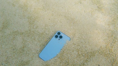 A close-up of a modern fashionable phone lying on bottom of the ocean with white sand. A fashionable blue phone got lost on the seabed with solar glare. Tests for water resistance of the new phone.