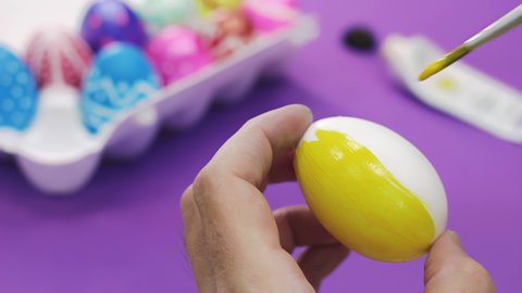 Happy Easter Close Up. A white man paints an Easter egg with yellow paint on a purple tabletop. Easter Holidays preparation theme. Selective focus