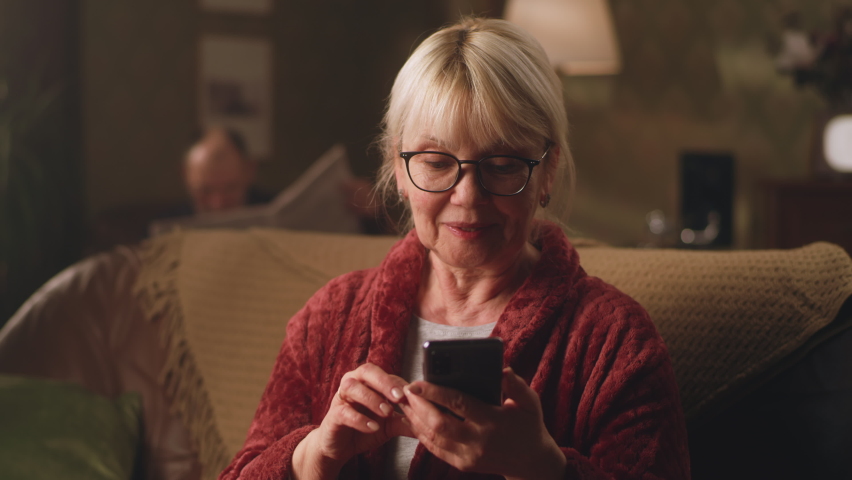 Excited senior woman staring at camera and rejoicing at unexpected message on smartphone while sitting on sofa at home Royalty-Free Stock Footage #1088689295