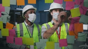Diverse architects in safety mask and hardhat discuss and write on glass board with sticky notes working together in office - Powered by Shutterstock - Get 15% off with code: PIKWIZARD15