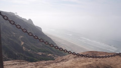 Steep unstable cliff, rock or bluff, foggy weather, California coast erosion, USA. Torrey Pines vista, eroded crag overlook viewpoint, ocean waves from above. Chain railing for safety trekking, danger