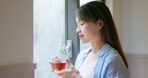asian smiling young woman is drinking tea while looking through window on a rainy day at home