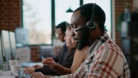 Male worker answering telework phone at call center, using audio headset and microphone. Young man working at customer care in friendly office. Handheld shot. - Powered by Shutterstock - Get 15% off with code: PIKWIZARD15
