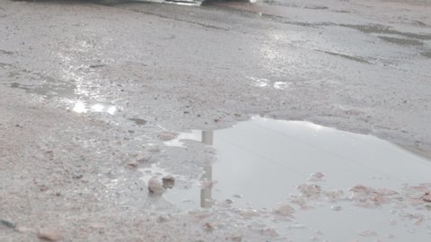 Water-filled potholes on an asphalt road with passing cars. Rainy weather. The car drives through puddles on broken asphalt roads after rain during the daytime. Raindrops and cars on the road.