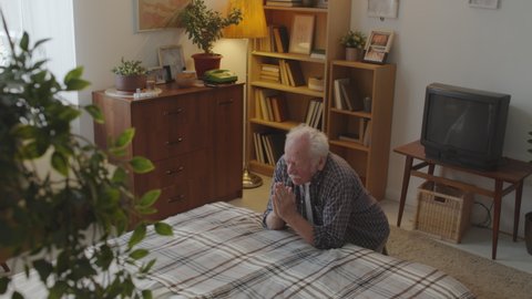 High angle tilting down of senior Caucasian man with white mustache praying in his room, kneeling by bed in evening
