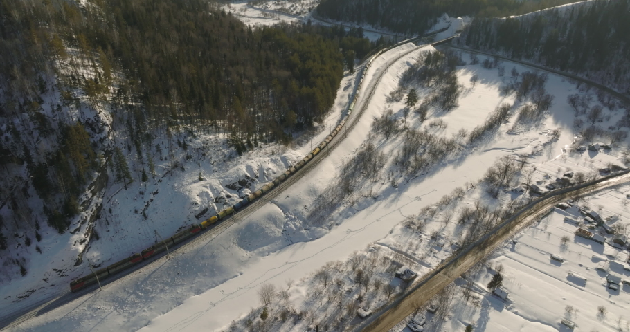 Freight long train carries with oil tank and petrol carriages an electric locomotive by Trans Siberian railways under the rock and near mountain river. Aerial drone wide view at winter sunny sunset Royalty-Free Stock Footage #1088699255