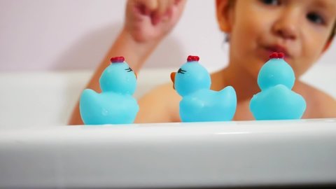 A little boy takes a blue rubber duckling out of three on the edge of a bath while washing