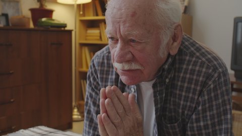 Medium close-up of retired Caucasian man with white mustache praying in his room, kneeling by bed in evening