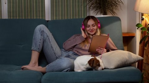 Attractive girl listens to music and watches video clips on a tablet as a dog in a home environment indoors Video de stock