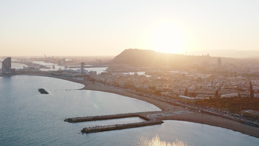 Wide angle panoramic aerial view of Barcelona shoreline. Sun setting behind the hill shine on Barceloneta Beach golden sand create glare on calm sea surface at cozy bay. Hotel W is seeing on the left | Shutterstock HD Video #1088704673