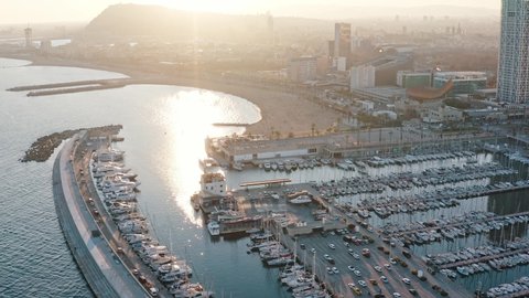 Panning shot from air on Port Olimpic, Barcelona crowded with yachts sailing ships in lights of setting sun. Hotel Arts building is near and Hotel W silhouette similar to a sail can be seeing far away