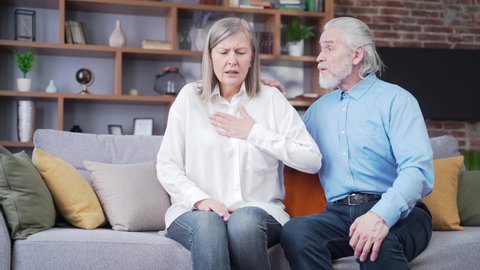Senior mature woman helping her husband with chest pain. Old man feeling bad pain disease. Wife supporting husband - Elderly couple suffering from symptoms heart attack ache. sitting at home indoors