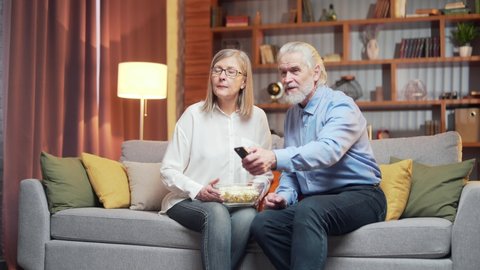 grey-haired older couple at home choose a television show to watch and switch channels with a TV remote control. elderly mature family husband and wife cannot choose a show. watching news or film