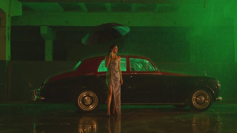 Beautiful Elegant Young Woman Standing and holding umbrella near old retro classic car. Girl with umbrella stands in street under the rain . Old fashioned luxury style concept . Raining outside 
