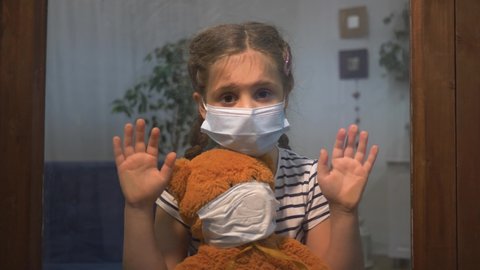 Protection of children, medical. Sad girl in protective medical mask at window. girl is alone at home with toy. sad child in protective mask in hospital looks out window. child is isolated at home.
