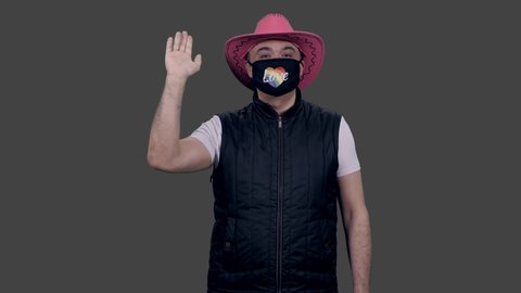 Gay person in anti covid lgbt mask and pink cowboy hat showing solidarity gesture and waving hand, 4k footage with alpha transparency channel isolated on gray background