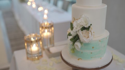 Handheld of a three layer wedding cake with flowers near the dinning tables decorated with golden candles as center pieces in indoor venue