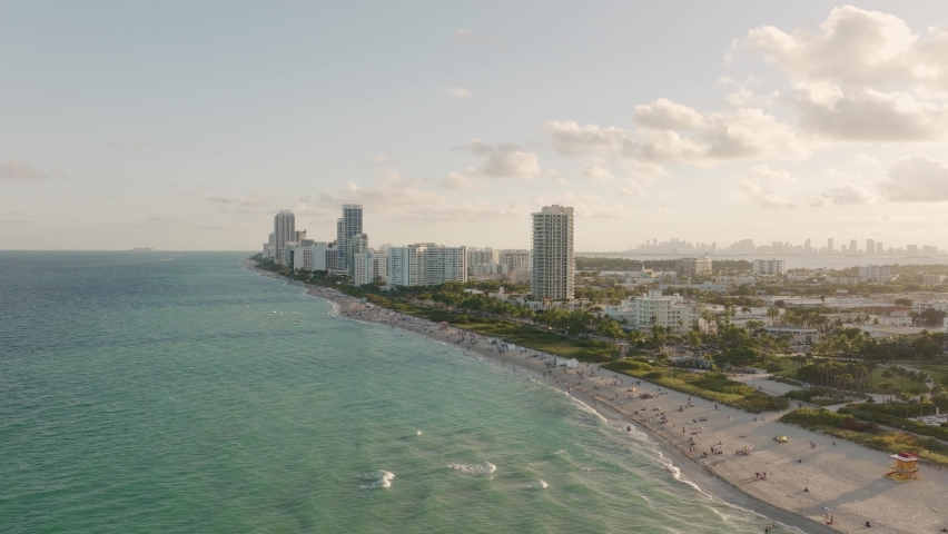 Aerial trucking of people relaxing in sand shore near turquoise sea in Miami Beach, resorts and apartment buildings in background at golden hour, Florida, USA