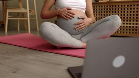 Close up pregnant woman sitting on mat in front of laptop stroking her big belly