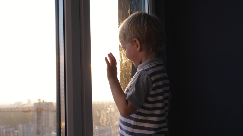 Little cheerful blond kid stands at window in high rise building. Looks at street exploring world with interest and curiosity. Against rays of sunshine at sunset. Child development and home games