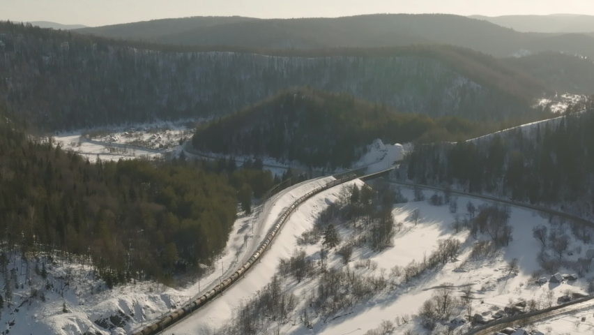 Freight long train carries with oil tank and petrol carriages an electric locomotive by Trans Siberian railways under the rock and near mountain river. Aerial drone wide view at winter sunny sunset Royalty-Free Stock Footage #1088723487