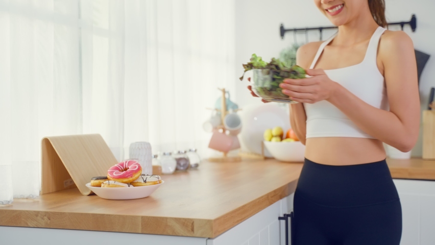 Asian beautiful sport woman choose to eat green salad instead of donut. Attractive active young girl feel happy and enjoy vegetables healthy foods for health after exercise workout in kitchen at home.