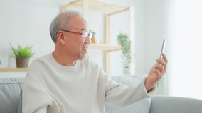 Asian senior older man use mobile phone for video call in living room. Attractive elderly mature male feel happy while sit on sofa, using smartphone talk to video conference call with family in house.