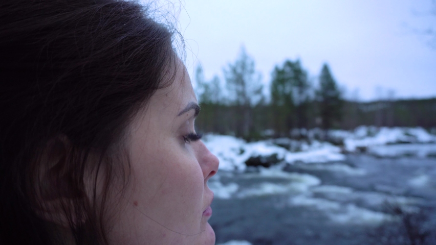 A beautiful girl in a blue dress at a winter river looks into the distance and smiling. Woman in the winter forest near the river between the rocks. Portrait of a brunette in a snowy forest	