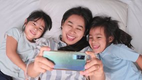 Happy Asian family enjoying with smartphone at cozy home. Smiling mother and cute daughters using phone, Watch video games or cartoons online in bed at home. Good time at home
